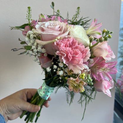 A hand holding a Prom bouquet of pink and white flowers, including roses, carnations, alstroemeria, and baby's breath, wrapped in a green and Rhinestone ribbon against a plain light gray wall in Florence, Kentucky designed by Flair Flower Bar