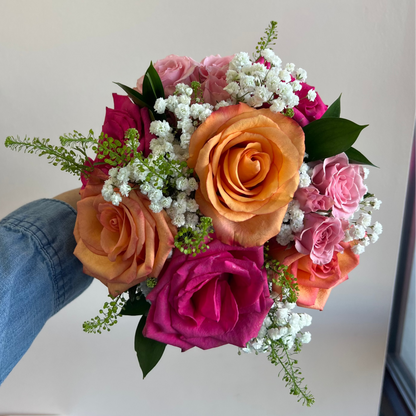 A hand holding a vibrant floral bouquet featuring orange and pink roses, magenta blooms, green foliage, and white baby's breath against a plain light background.