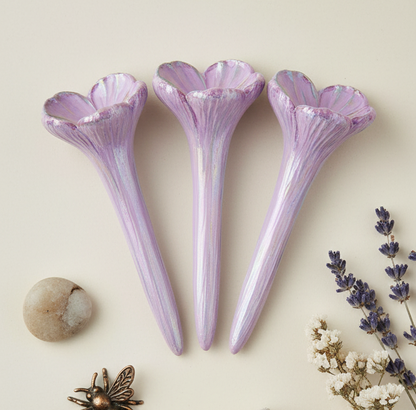 Three purple ceramic flower-shaped pollinator spikes on a light background with dried flowers and a stone.