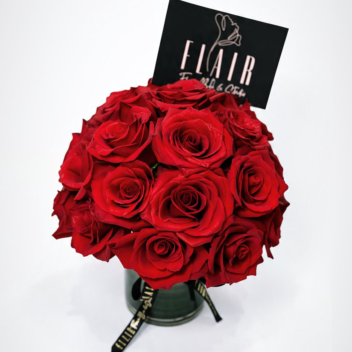 A round bouquet of red roses in a clear vase, featuring a black card with the brand name 'FLAIR' and a black ribbon with gold lettering, set against a plain white background.