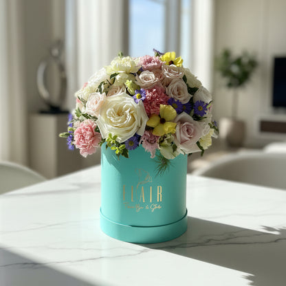 A vibrant, multicolored floral arrangement featuring white, pink, yellow, and purple flowers in a round turquoise box branded 'FLAIR Flower Box & Studio'. The box sits on a white countertop with a softly blurred indoor background.