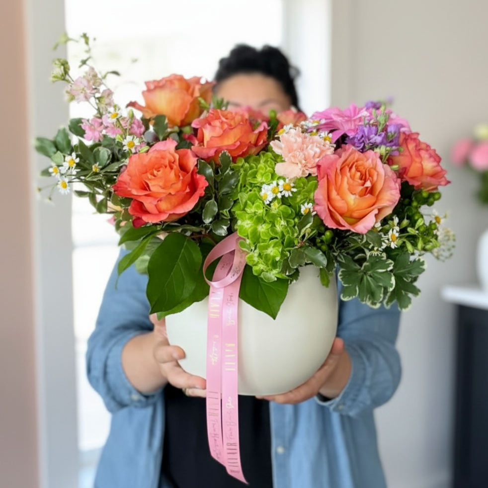A person in a blue denim shirt holds a large white round planter filled with a colorful floral arrangement featuring orange roses, pink hydrangeas, green foliage, and a pink ribbon, set against a warm indoor background. Designed by Flair Flower Bar & Studio in Florence, Kentucky