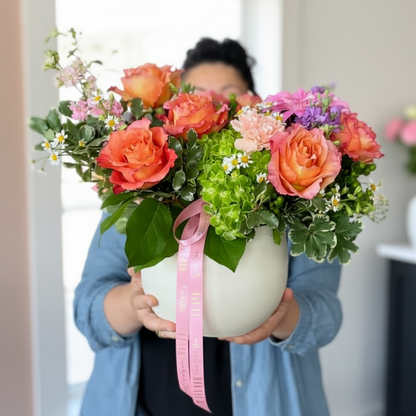 A person in a blue denim shirt holds a large white round planter filled with a colorful floral arrangement featuring orange roses, pink hydrangeas, green foliage, and a pink ribbon, set against a warm indoor background. Designed by Flair Flower Bar & Studio in Florence, Kentucky