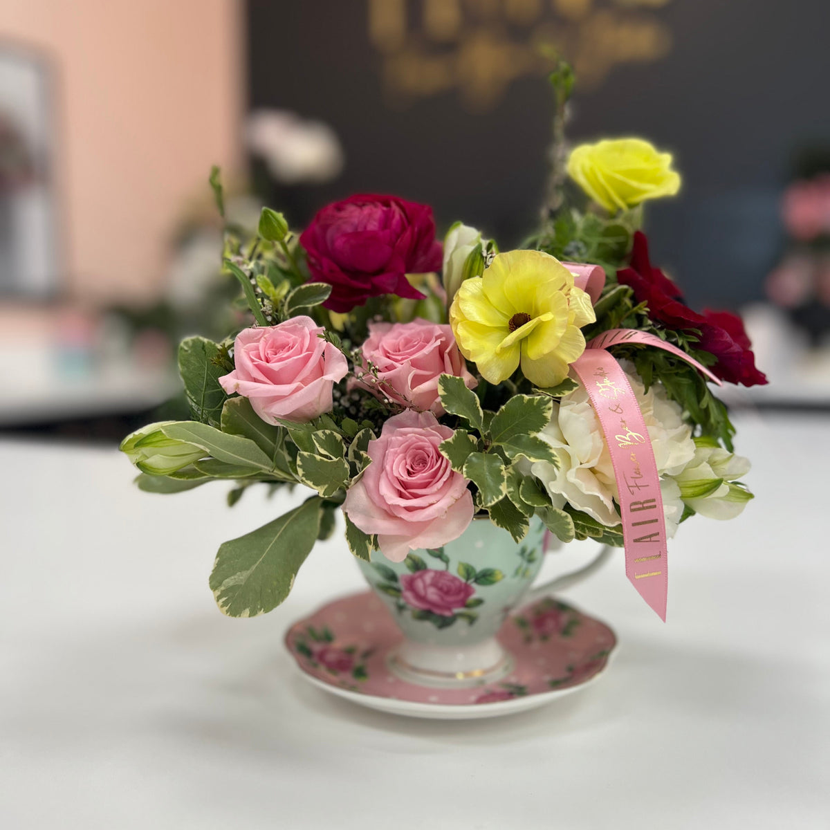 A floral arrangement in a vintage-style floral teacup and saucer, featuring pink and yellow flowers with green leaves, set on a white table with a blurred background.