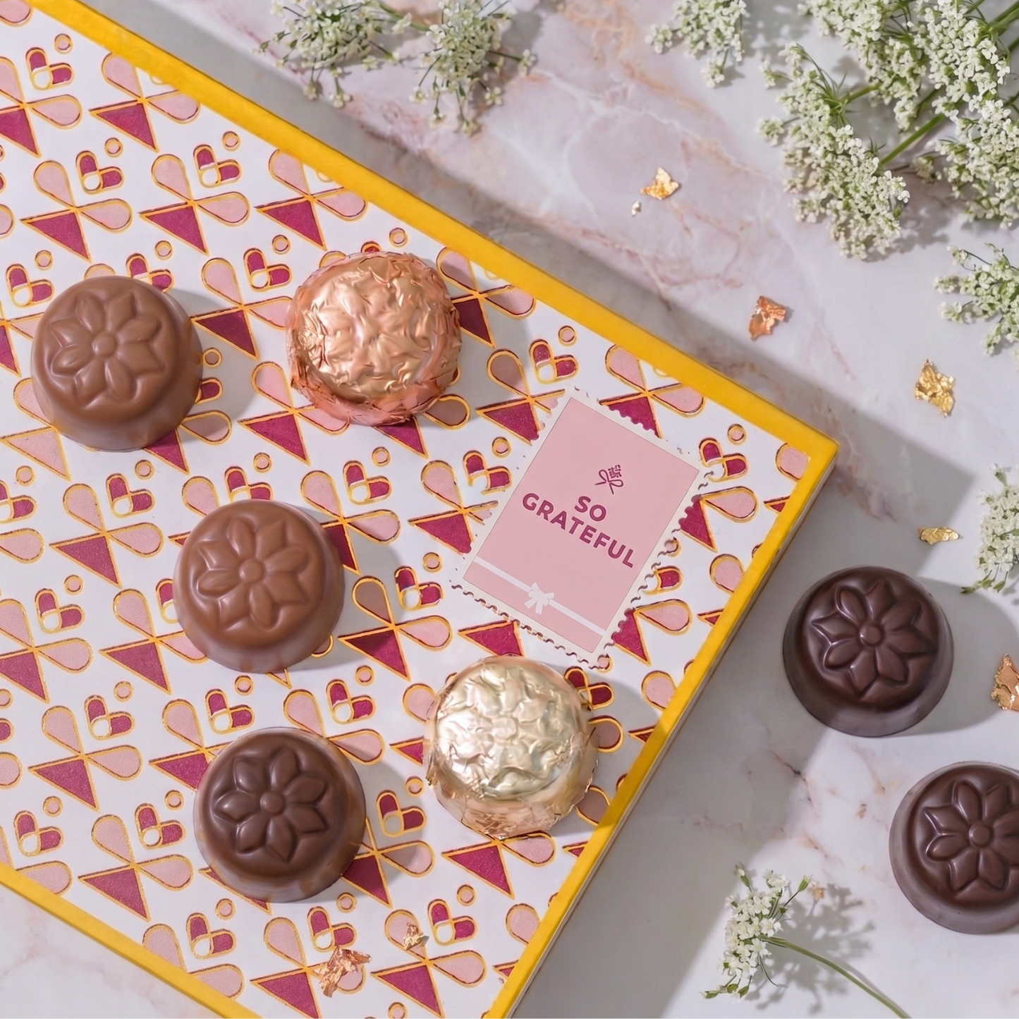 Chocolate truffles on a decorative box with 'So Grateful' card, surrounded by flowers.