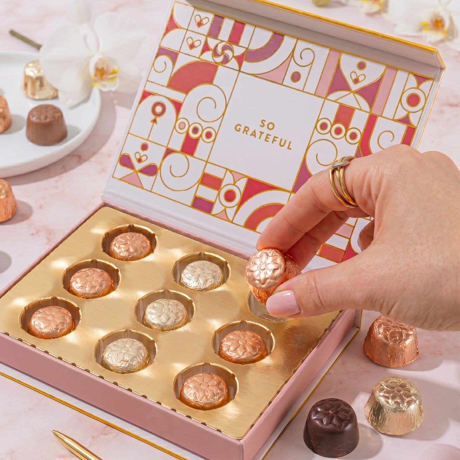 Hand reaching into a box of chocolates with a decorative cover on a light pink surface.
