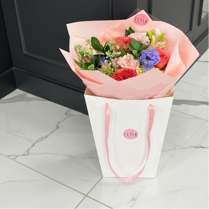Bouquet of flowers in a white paper bag with pink handles on a wooden floor.