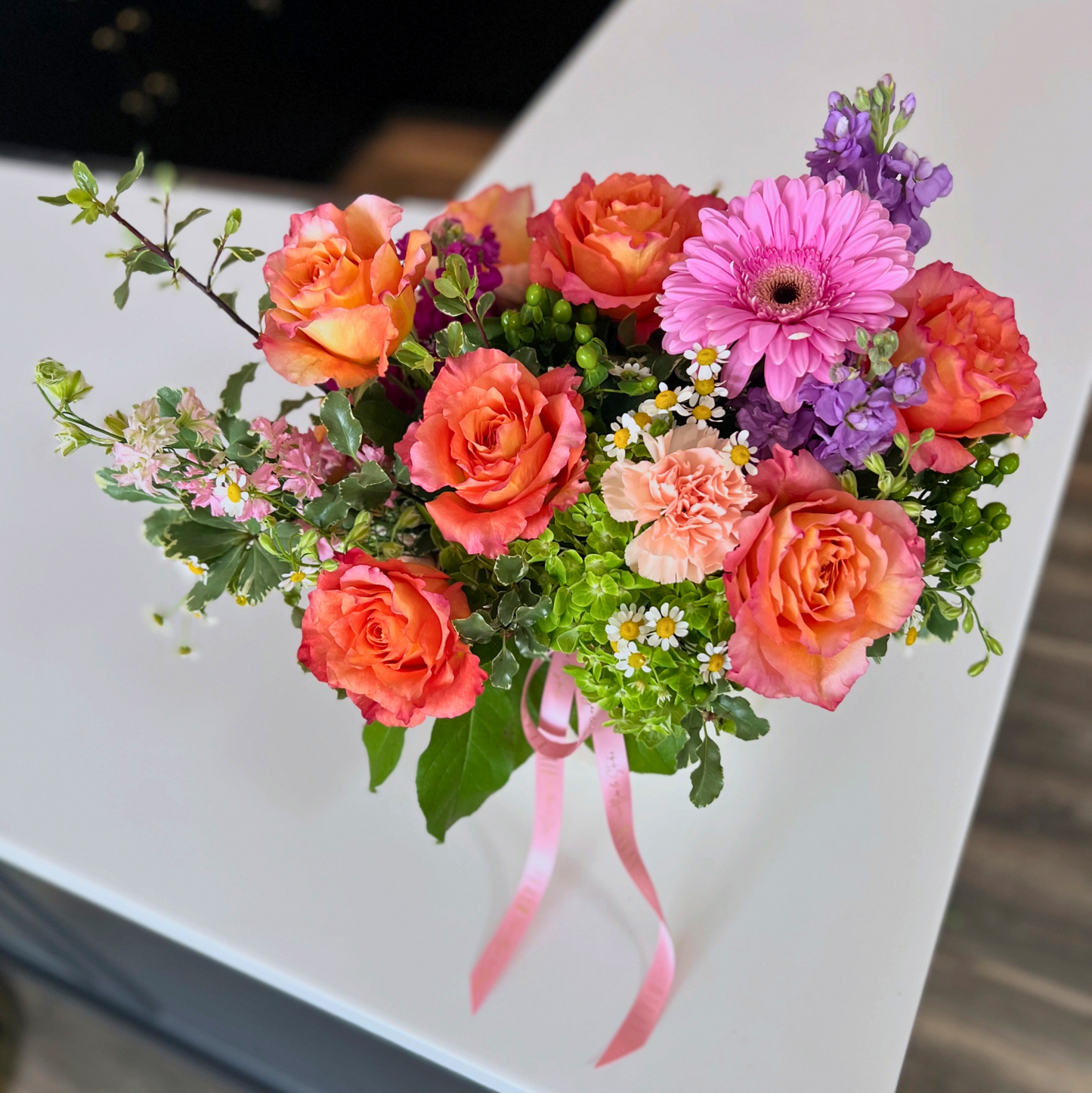 A vibrant floral bouquet featuring coral and pink roses, a pink gerbera daisy, purple flowers, and green foliage, tied with a pink ribbon and resting on a white tabletop by Flair Flower Bar & Studio in Florence, Kentucky