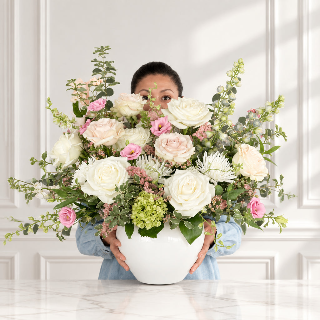 A person in a blue denim shirt holds a large white planter filled with a colorful floral arrangement featuring white and pink roses, green foliage, and small white blossoms inside a flower shop in Florence, Kentucky. 