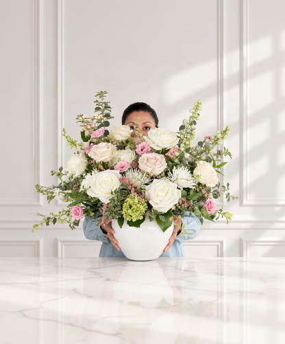 The Signature Garden Arrangement by Flair Flower Bar is a large white planter filled with a lush floral arrangement of white and pink roses, green foliage, and small hydrangea blooms, set against a white paneled wall with soft window shadows offering delivery throughout Cincinnati and Northern Kentucky.