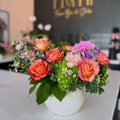 A vibrant floral arrangement featuring coral and pink roses, purple flowers, green hydrangeas, and small white daisies in a round white ceramic pot, set on a light gray counter with a blurred background showing a 'FLAIR' sign in Florence, Kentucky