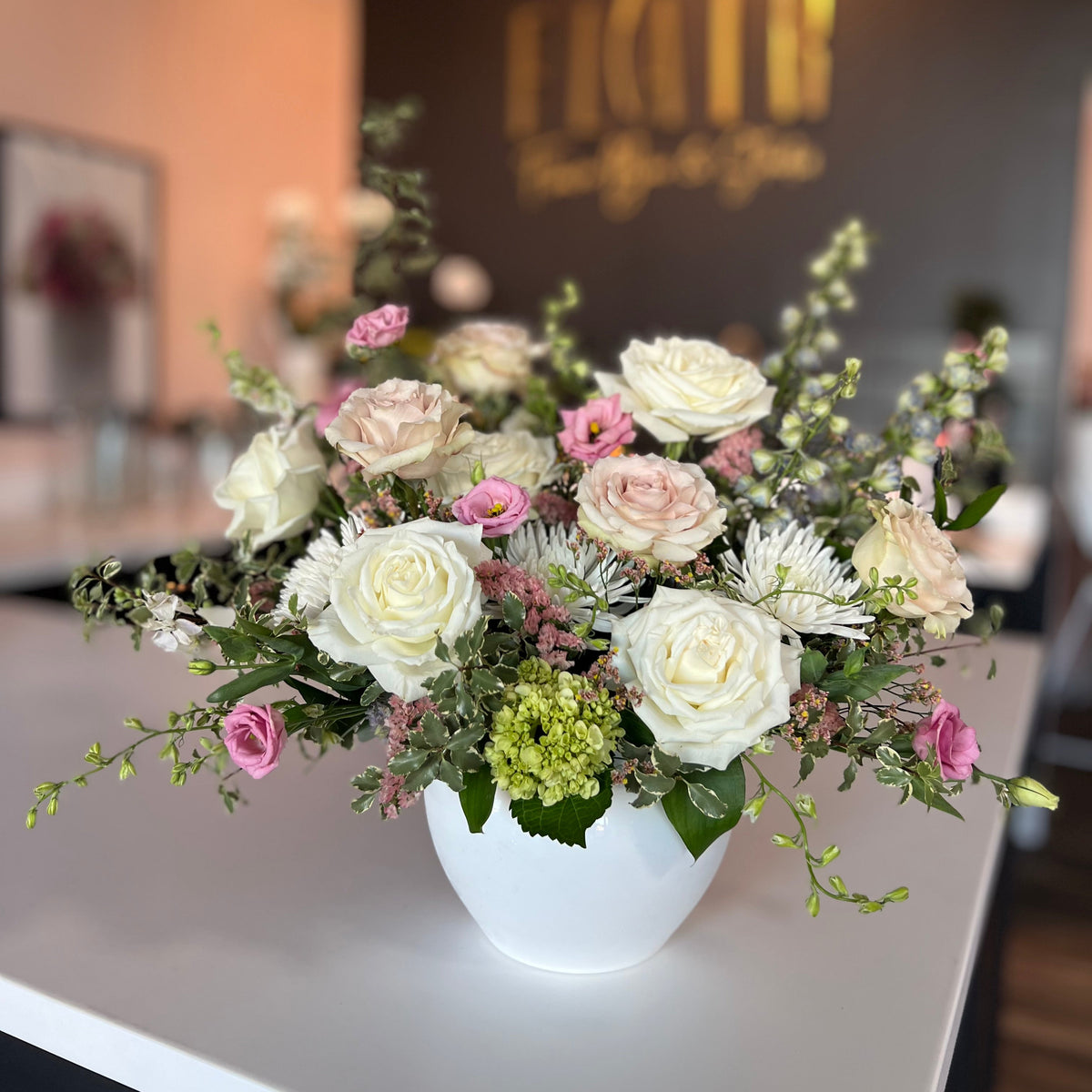 Flair Flower Bar's Signature Garden Flower Arrangement - A luxury floral arrangement of white and blush pink roses, green foliage, and small white daisy-like flowers in a white vase, placed on a white counter in a softly lit indoor setting. A gold-toned Flair Flower Bar logo is visible on a dark wall behind.
