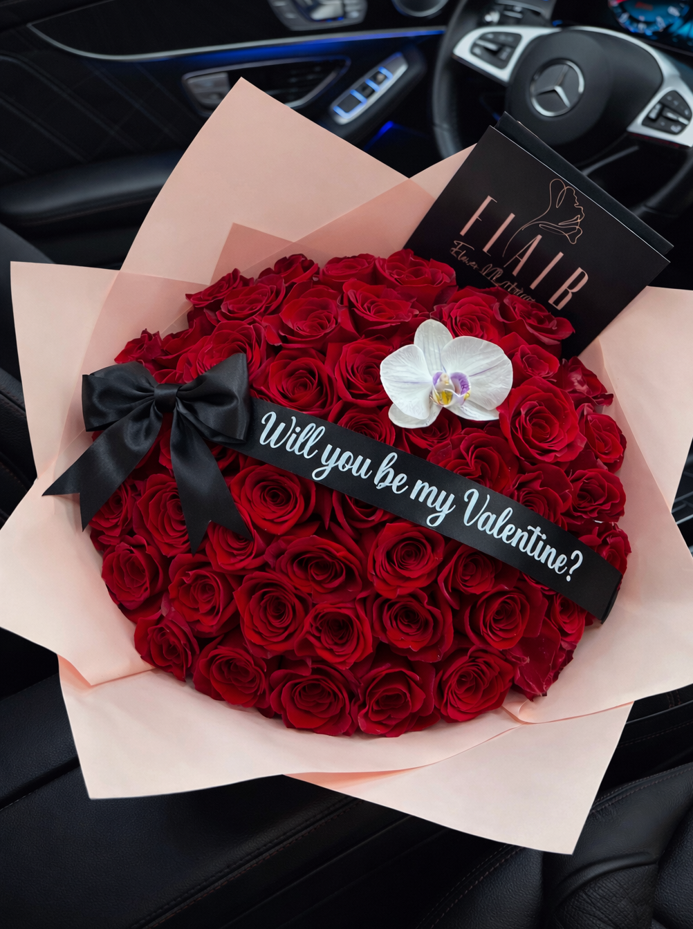Heart-shaped bouquet of red roses with a black bow and 'Will you be my Valentine?' message, placed on a car dashboard.
