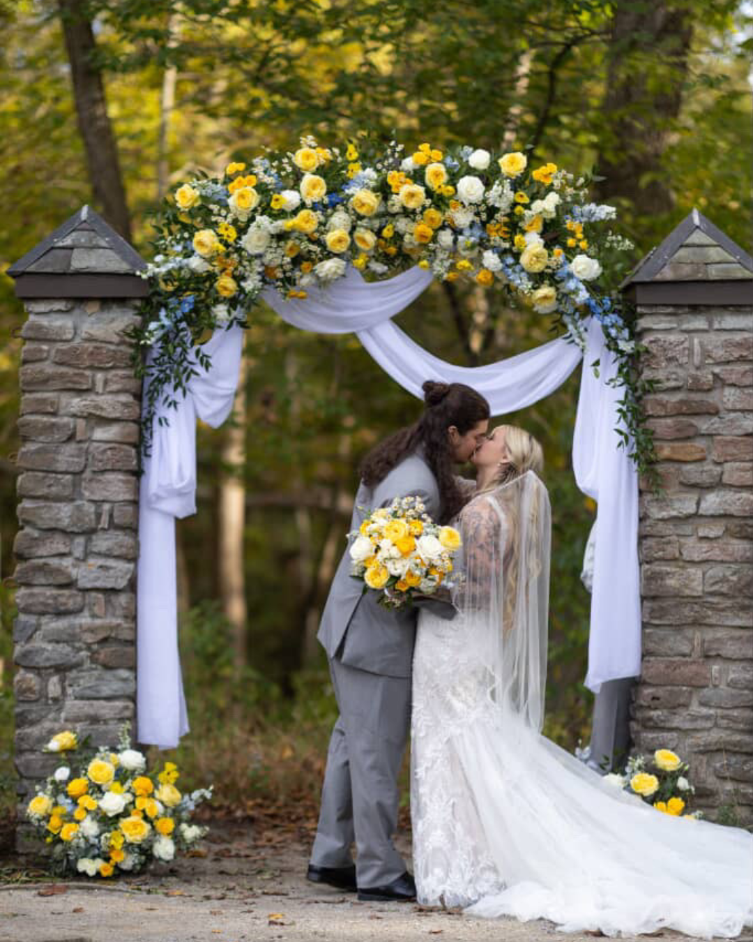 Wedding arch Cincinnati Nature Center_Groesbeck Estate Couple kissing under a floral archway with yellow and white flowers, surrounded by greenery.